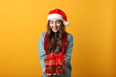 Photo of Happy young woman with tinsel, Santa hat and gift box on orange background