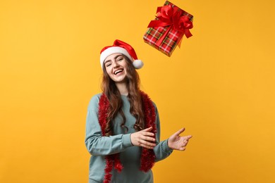 Photo of Happy young woman with tinsel and Santa hat throwing gift box on orange background