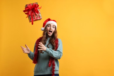 Photo of Emotional young woman with tinsel and Santa hat throwing gift box on orange background. Space for text