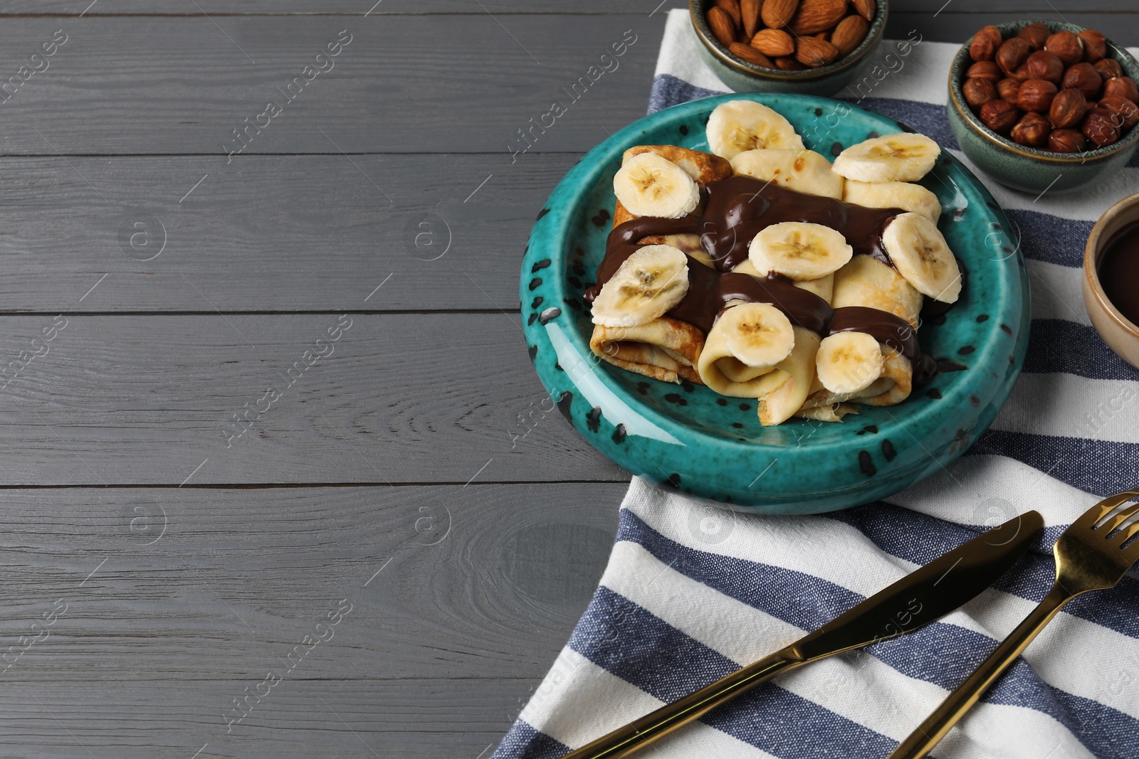 Photo of Delicious crepes with banana, nuts and chocolate paste on grey wooden table, space for text