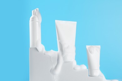 Photo of Tubes and bottle of cosmetic products with foam on podium against light blue background