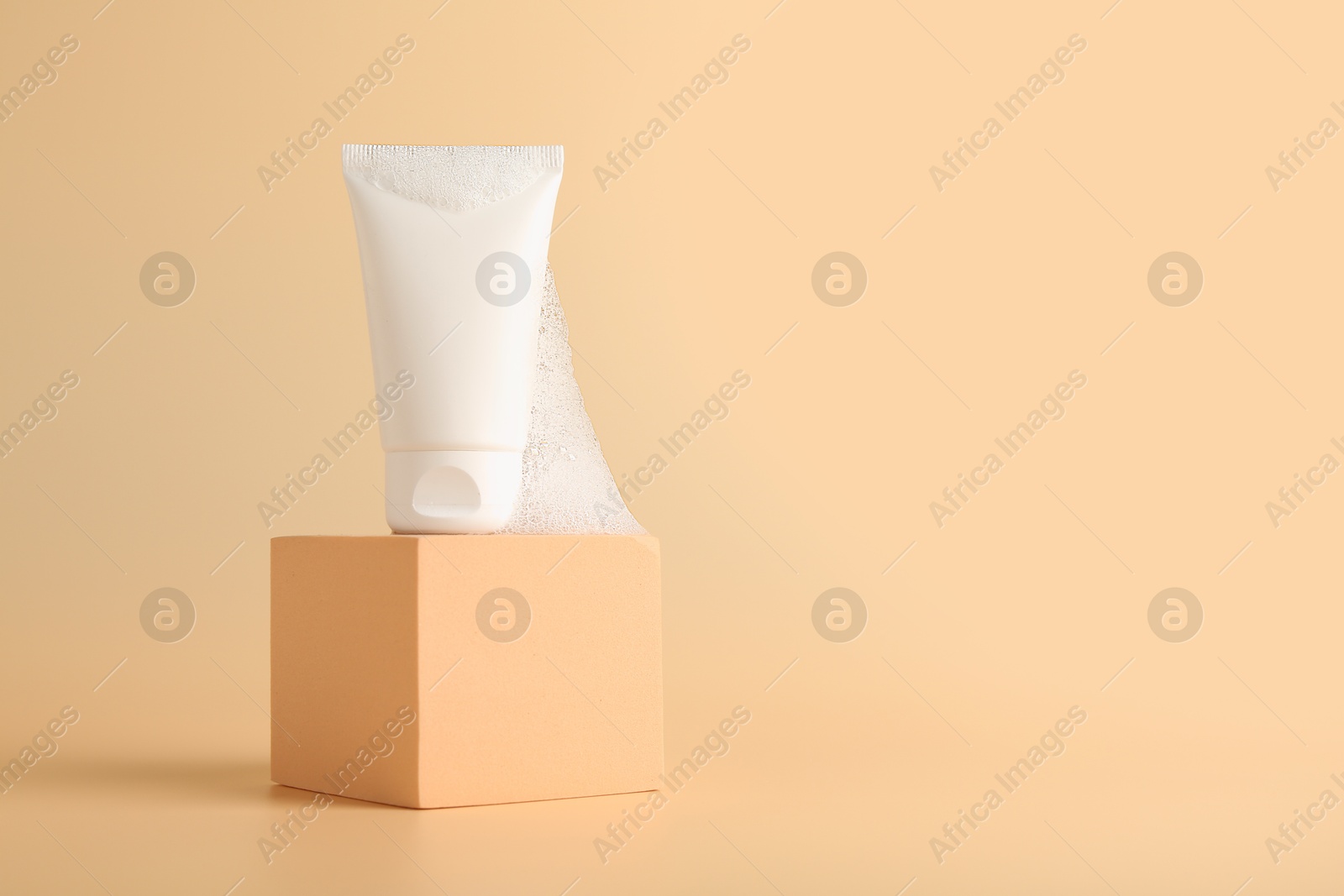 Photo of Tube of cosmetic product with foam on podium against beige background, space for text