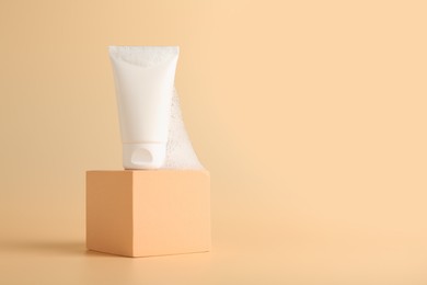 Photo of Tube of cosmetic product with foam on podium against beige background, space for text