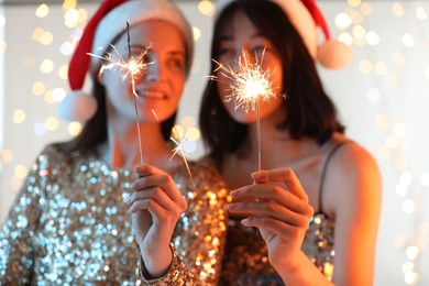 Photo of Women holding bright burning sparklers on background with blurred lights, selective focus