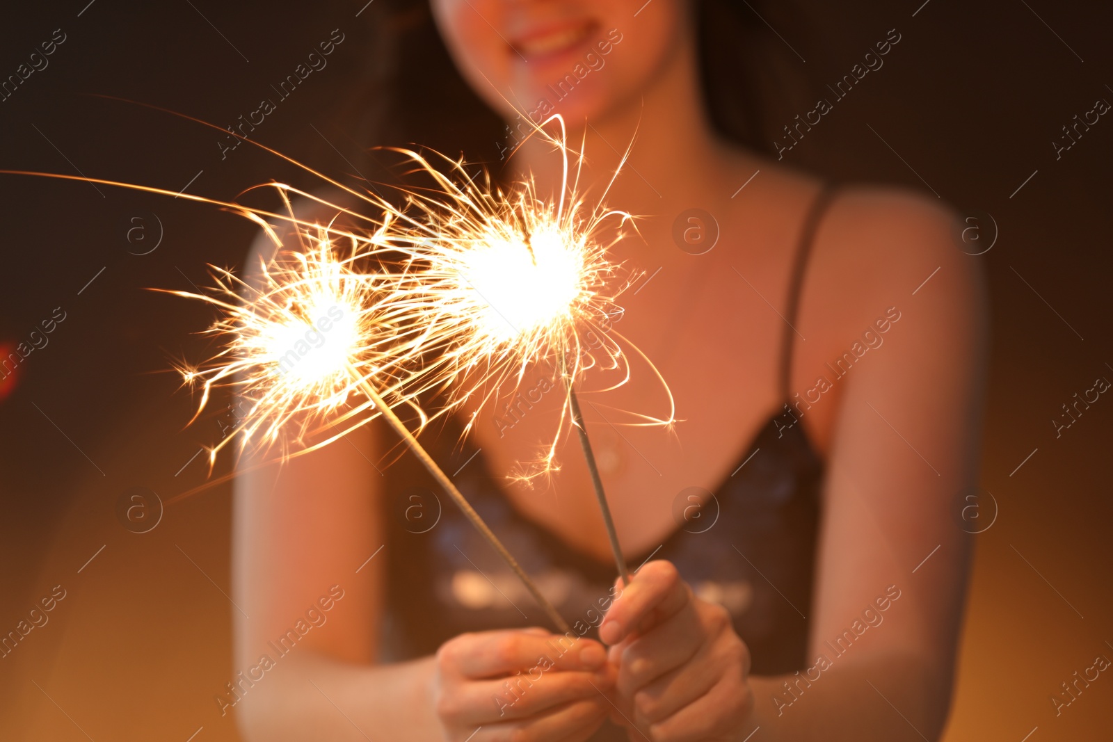 Photo of Woman with bright burning sparklers on dark golden background, selective focus