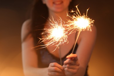 Photo of Woman with bright burning sparklers on dark golden background, selective focus