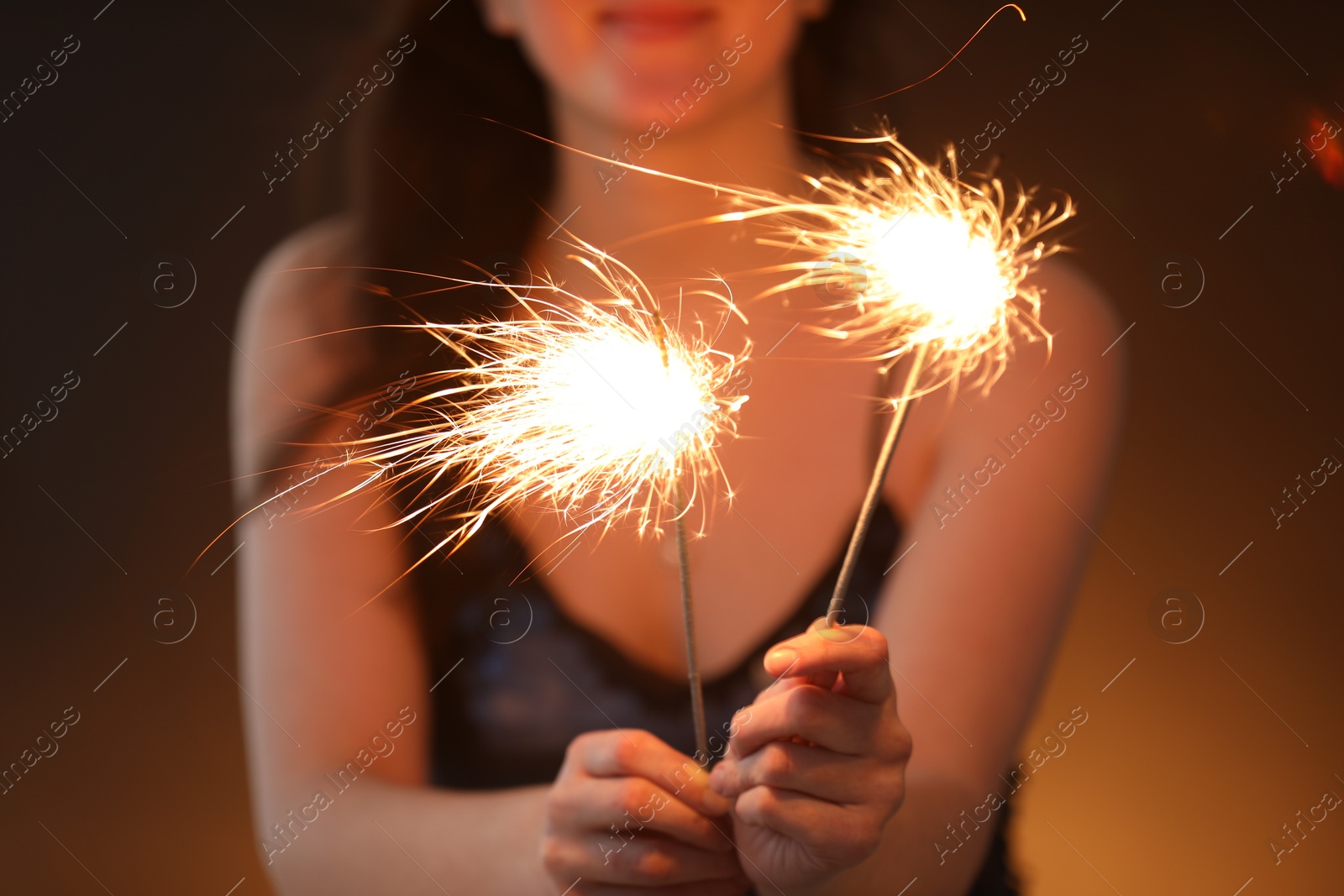 Photo of Woman with bright burning sparklers on dark golden background, selective focus