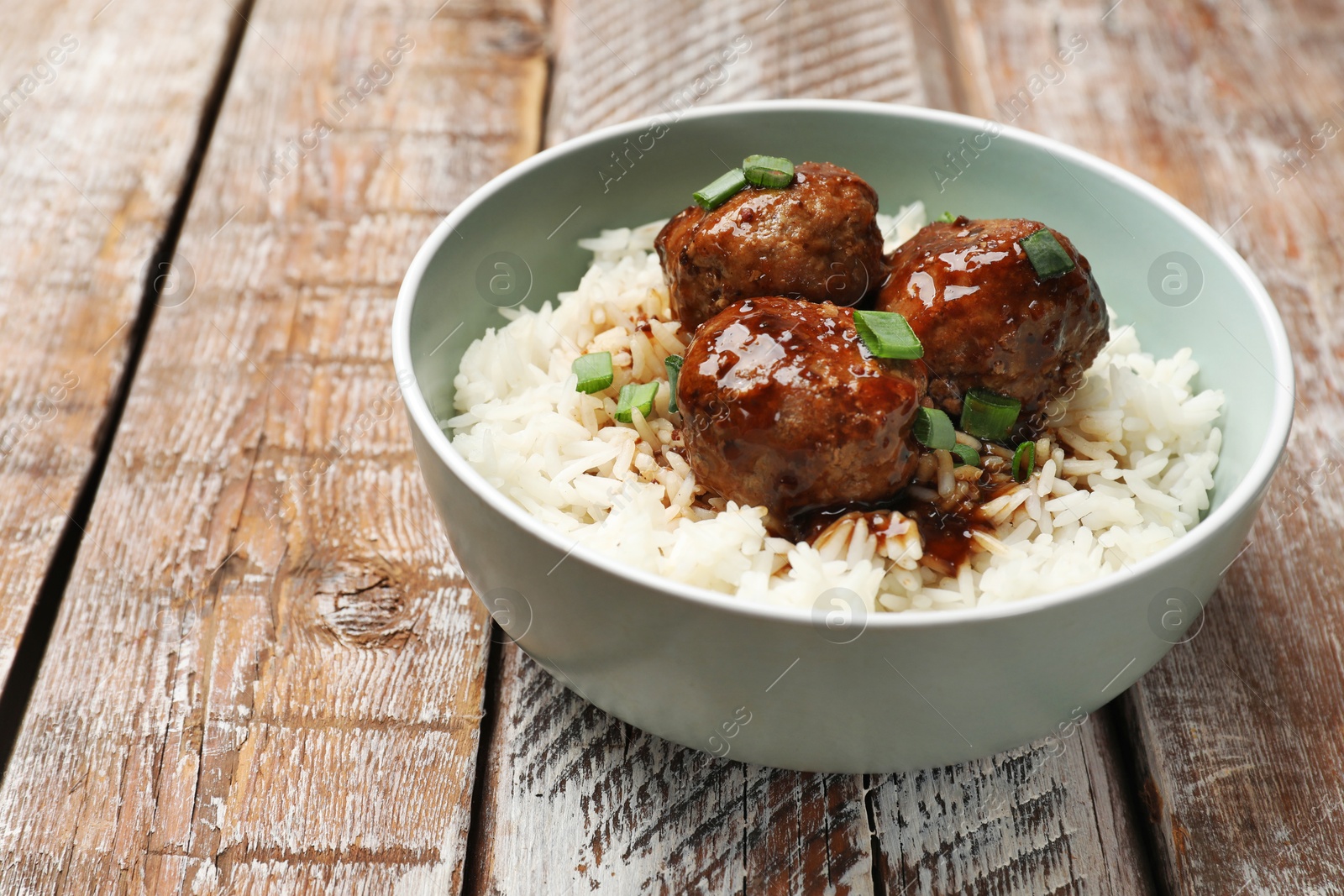 Photo of Delicious rice with meatballs, sauce and green onions in bowl on wooden table, closeup