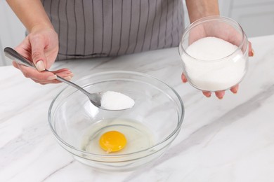 Photo of Making dough. Woman adding sugar into bowl with egg at white marble table, closeup