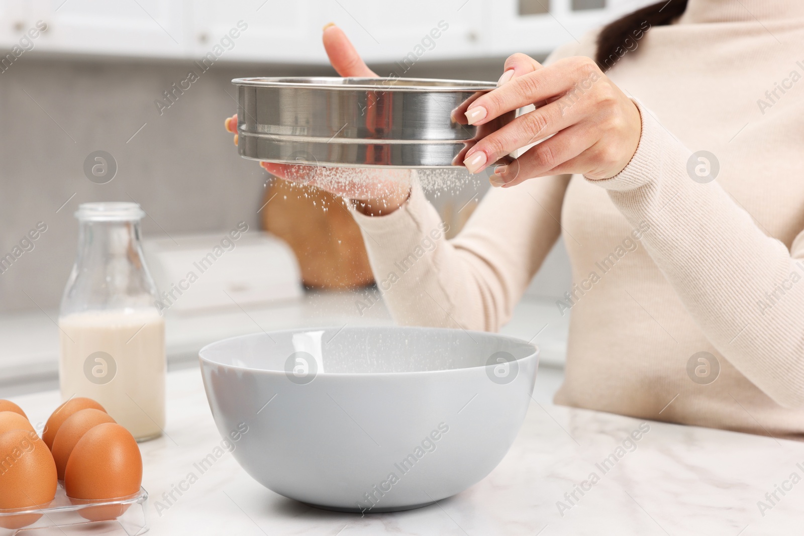 Photo of Making dough. Woman sifting flour into bowl at white table, closeup