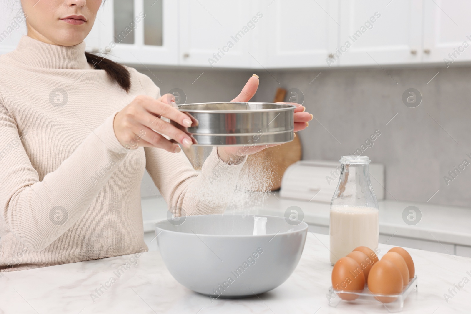 Photo of Making dough. Woman sifting flour into bowl at white table, closeup