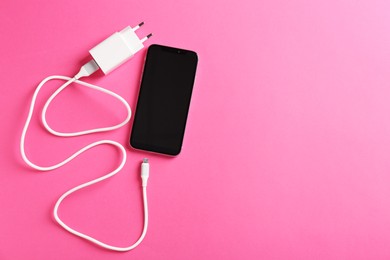 Photo of USB power adapter with charge cable and smartphone on pink background, flat lay. Space for text