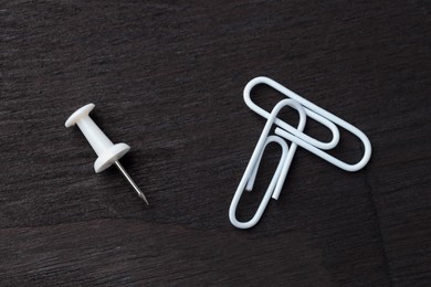 Two white paper clips and pin on black wooden table, flat lay