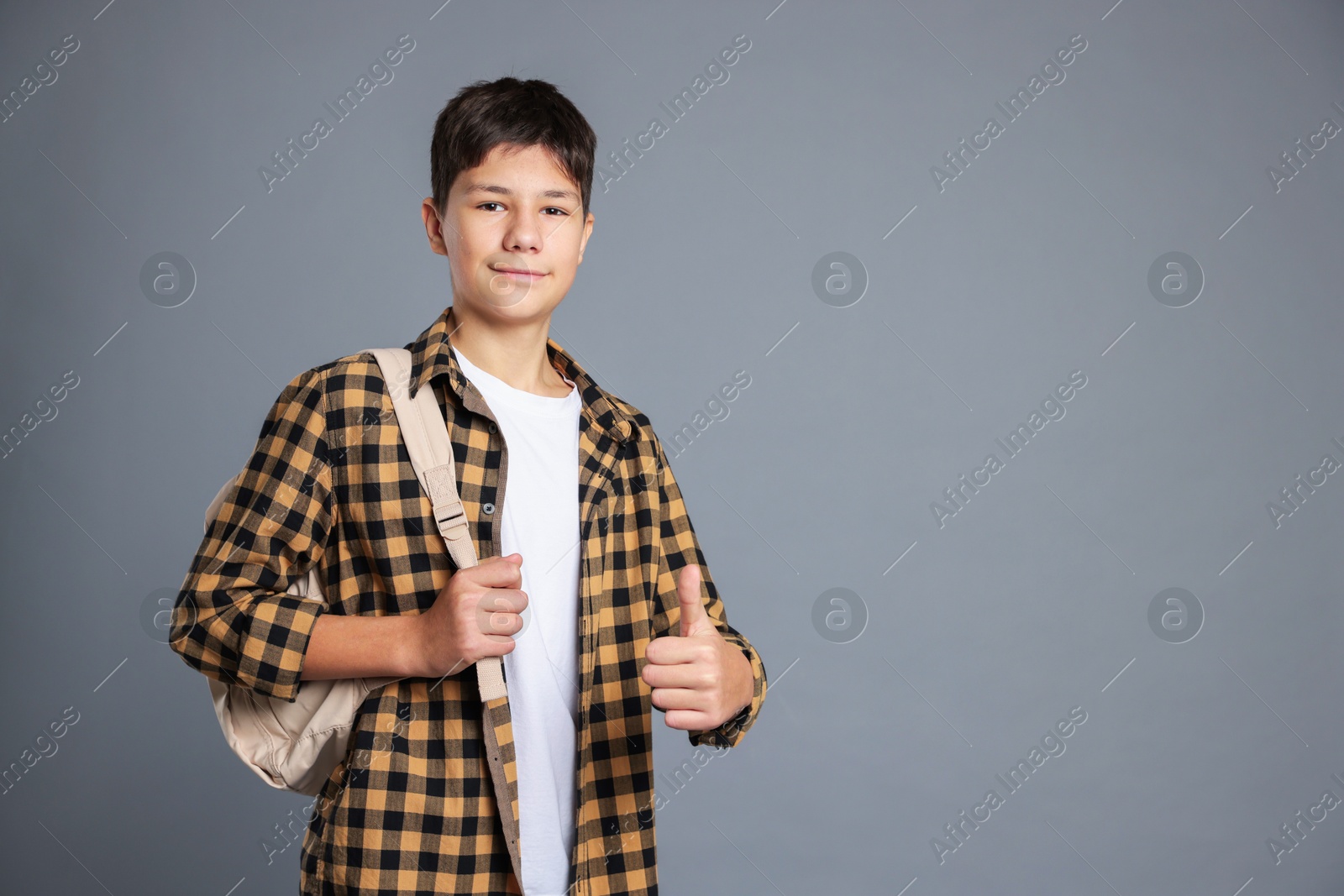 Photo of Portrait of teenage boy with backpack showing thumbs up on grey background, space for text