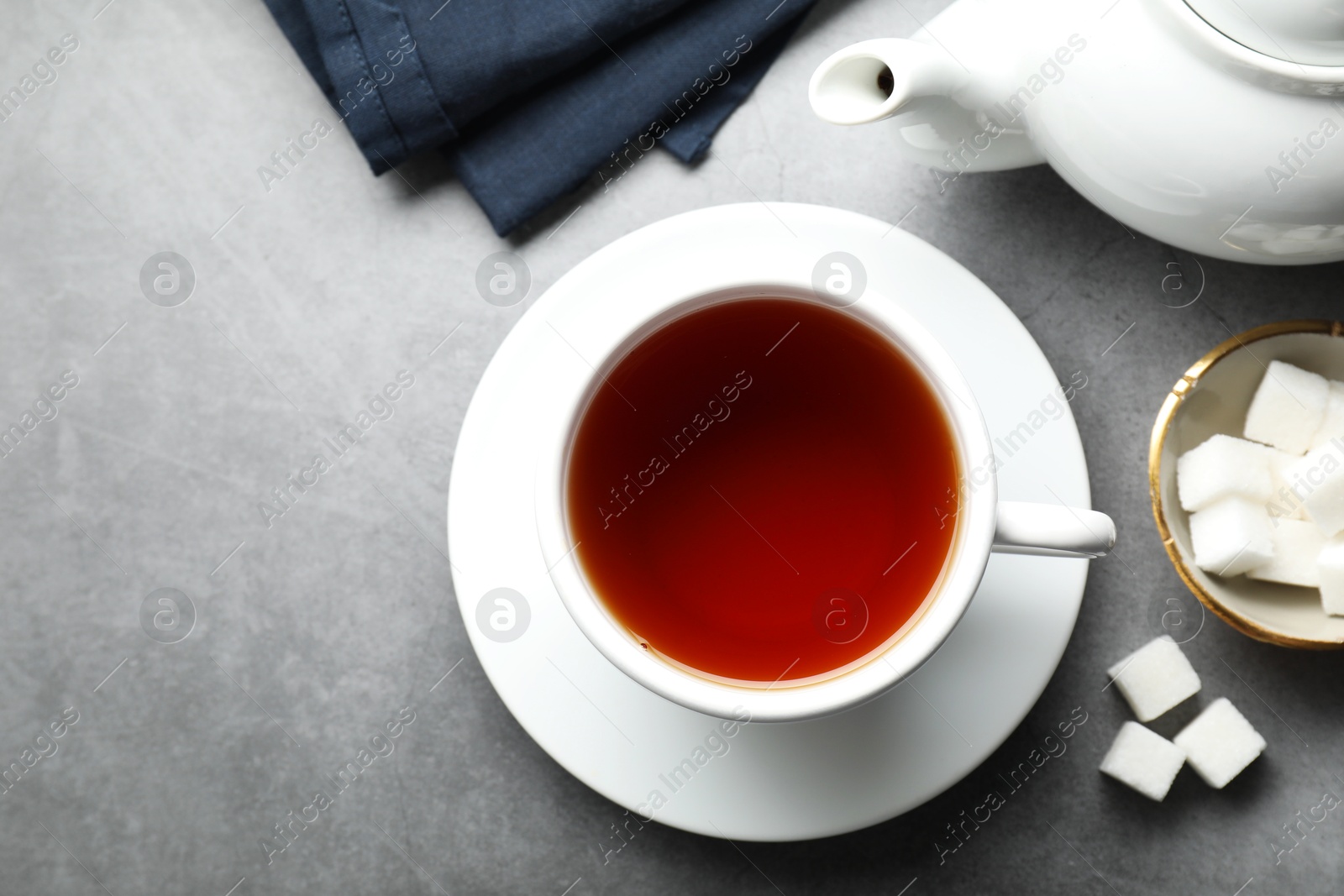 Photo of Refreshing black tea in cup, sugar cubes and teapot on grey textured table, flat lay. Space for text