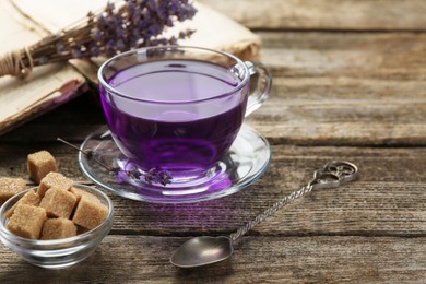 Photo of Aromatic lavender tea in glass cup, brown sugar, book, spoon and bunch of dry flowers on wooden table, closeup