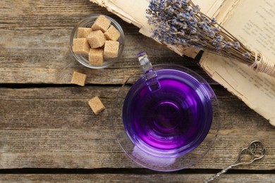 Photo of Aromatic lavender tea in cup, brown sugar, book, spoon and bunch of dry flowers on wooden table, flat lay. Space for text