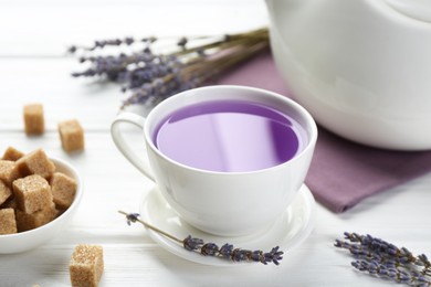 Photo of Aromatic lavender tea in cup, brown sugar and dry flowers on white wooden table, closeup