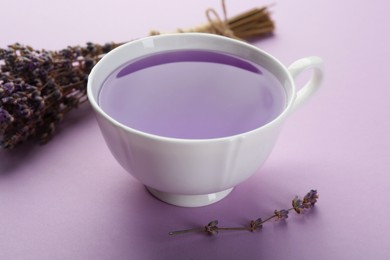 Photo of Aromatic lavender tea in cup and bunch of dry flowers on lilac background, closeup