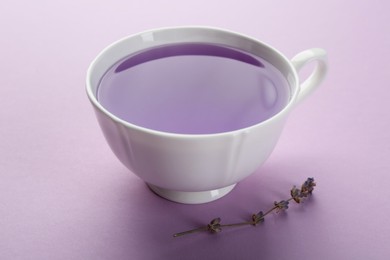 Photo of Aromatic lavender tea in cup and dry flowers on lilac background, closeup