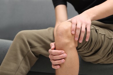 Photo of Young man suffering from pain in knee on sofa at home, closeup