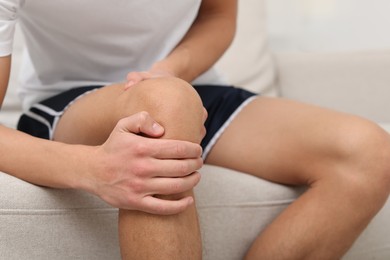 Photo of Young man suffering from pain in knee on sofa at home, closeup