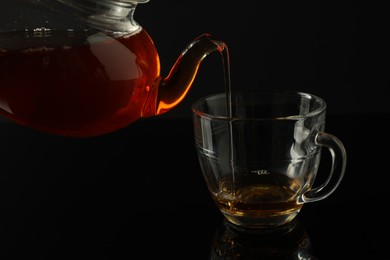 Photo of Pouring freshly brewed tea from teapot into glass cup on black table, closeup