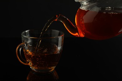 Photo of Pouring freshly brewed tea from teapot into glass cup on black table, closeup
