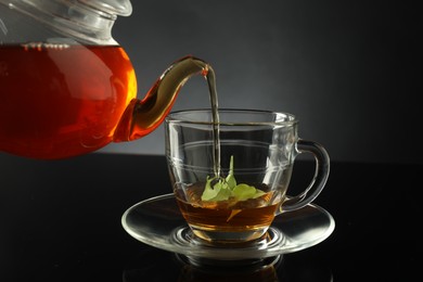 Photo of Pouring freshly brewed tea from teapot into glass cup on black table, closeup