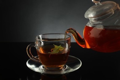 Photo of Pouring freshly brewed tea from teapot into glass cup on black table, closeup
