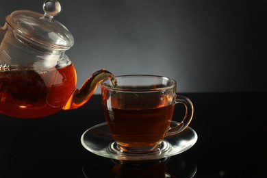 Photo of Pouring freshly brewed tea from teapot into glass cup on black table, closeup