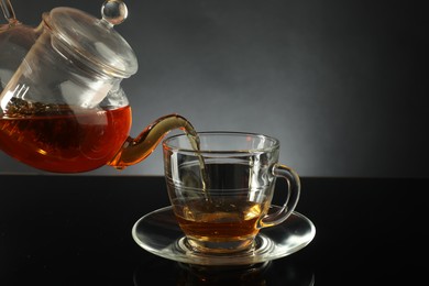 Photo of Pouring freshly brewed tea from teapot into glass cup on black table, closeup