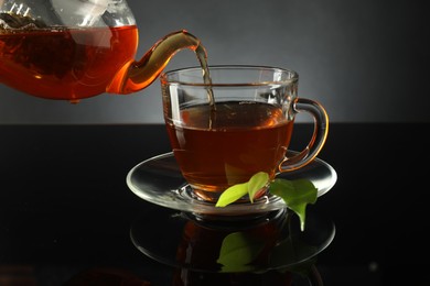 Photo of Pouring freshly brewed tea from teapot into glass cup on black table, closeup