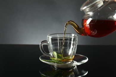 Photo of Pouring freshly brewed tea from teapot into cup at dark mirror table, closeup