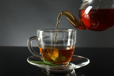 Photo of Pouring freshly brewed tea from teapot into cup at dark mirror table, closeup