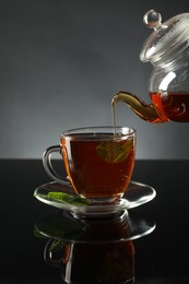 Photo of Pouring freshly brewed tea from teapot into cup at dark mirror table, closeup