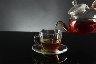 Photo of Pouring freshly brewed tea from teapot into cup at dark mirror table, closeup