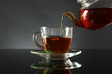 Photo of Pouring freshly brewed tea from teapot into cup at dark mirror table, closeup