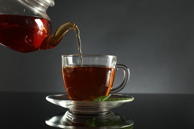 Photo of Pouring freshly brewed tea from teapot into cup at dark mirror table, closeup