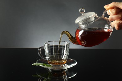 Photo of Woman pouring freshly brewed tea from teapot into cup at dark mirror table, closeup