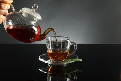 Photo of Woman pouring freshly brewed tea from teapot into cup at dark mirror table, closeup