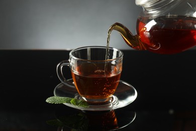 Photo of Pouring freshly brewed tea from teapot into cup at dark mirror table, closeup