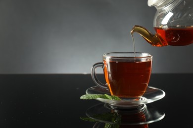 Photo of Pouring freshly brewed tea from teapot into cup at dark mirror table, closeup. Space for text