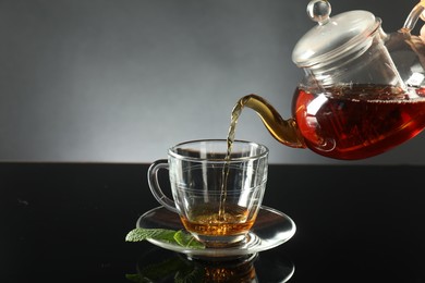 Photo of Pouring freshly brewed tea from teapot into cup at dark mirror table, closeup