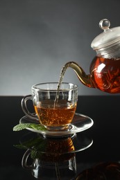 Photo of Pouring freshly brewed tea from teapot into cup at dark mirror table, closeup