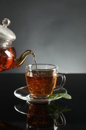 Photo of Pouring freshly brewed tea from teapot into cup at dark mirror table, closeup