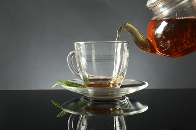Photo of Pouring freshly brewed tea from teapot into cup at dark mirror table, closeup