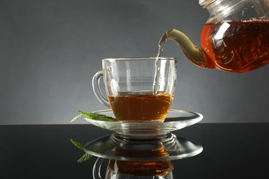 Photo of Pouring freshly brewed tea from teapot into cup at dark mirror table, closeup