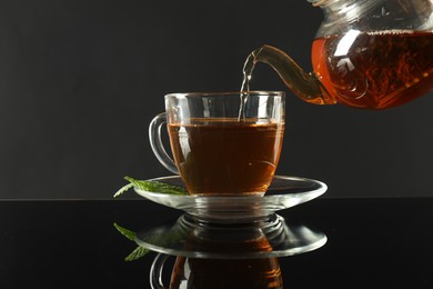 Photo of Pouring freshly brewed tea from teapot into cup at dark mirror table, closeup