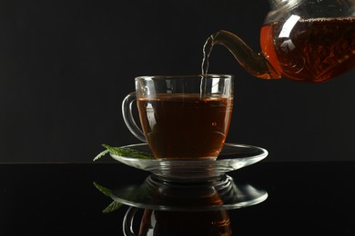 Photo of Pouring freshly brewed tea from teapot into cup at dark mirror table, closeup